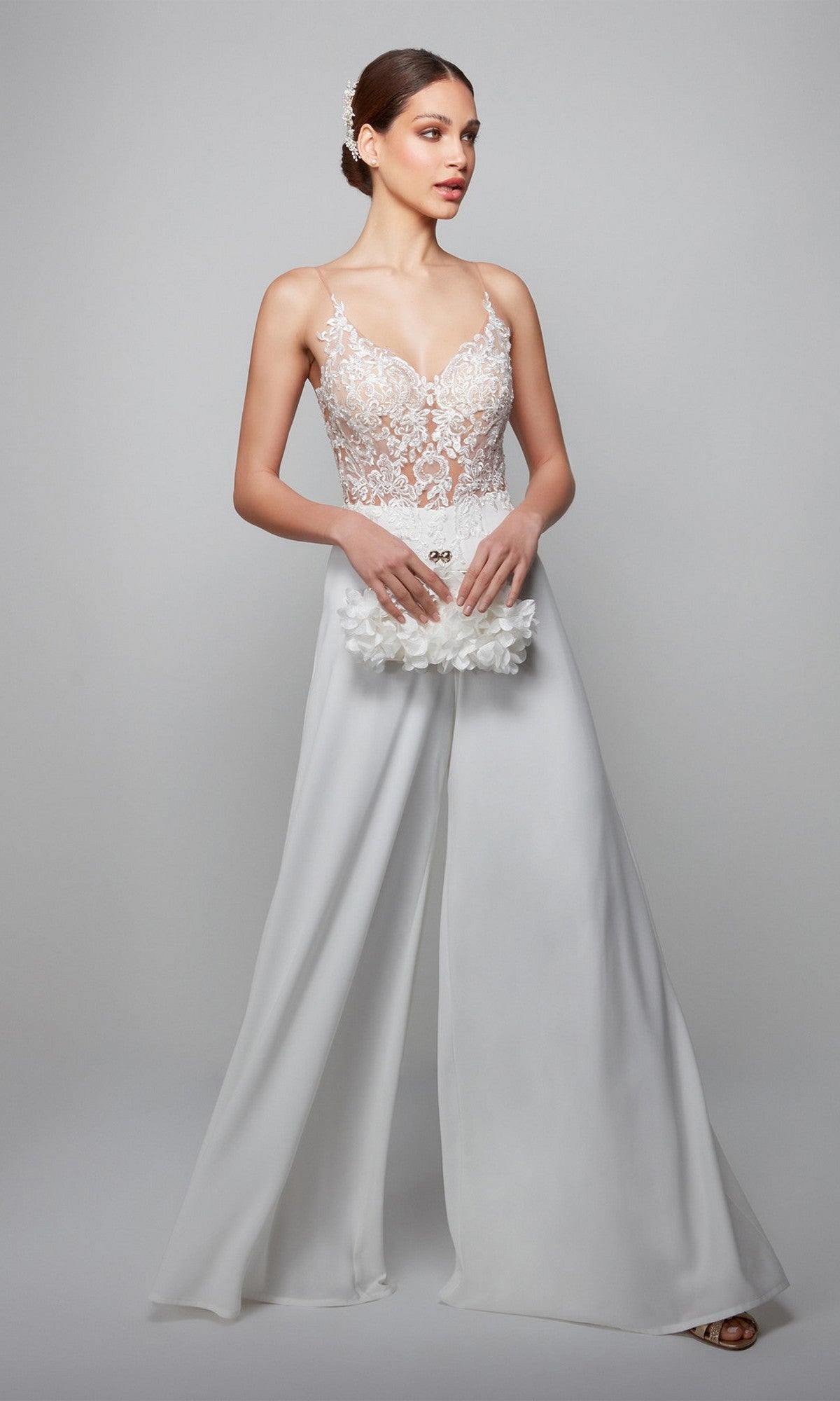 A woman wearing the Alyce 70017 Long White Jumpsuit with a lace bodice and wide-leg pants holds a white floral clutch, standing against a plain gray background.